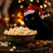 Black cat wearing a Santa hat sitting next to a bowl of popcorn with a festive background.