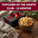 Bowl of popcorn next to a gift box with a red ribbon on a wooden table, with text 'Popcorn of the Month Club - 12 Months' at the top.
