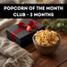 Bowl of popcorn next to a gift box with a red ribbon on a wooden surface, promotional text for 'Popcorn of the Month Club'.