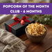 Bowl of popcorn next to a gift box with a red ribbon on a wooden surface, with text 'Popcorn of the Month Club - 6 Months' at the top.