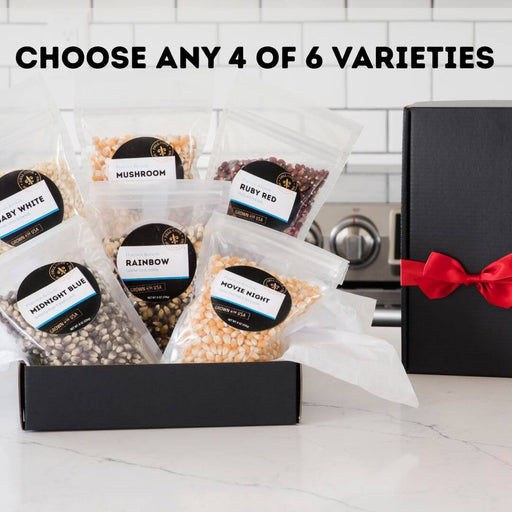 Six varieties of Dell Cove gourmet popcorn kernels displayed in resealable pouches beside a black gift box with red satin ribbon. Choose any four varieties.