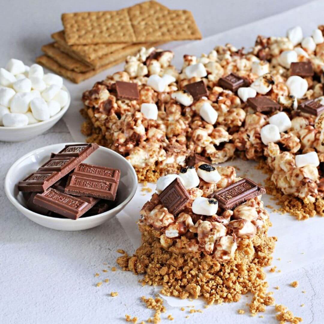 S'mores popcorn bar recipe with popped popcorn mixed with graham crackers, chocolate bars, and toasted marshmallows on a light surface.