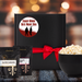 Black gift box with white label. Blood spilled and splattered on bottom half of label and a little across the top. The words Scary Movie Date Night Box in red are between the blood. Label shows a boy-boy zombie couple silhouette. Box is tied closed with red ribbon. Next to box is 2 popcorn seasoning pouches and a large bowl filled with popcorn. Dell Cove Spices