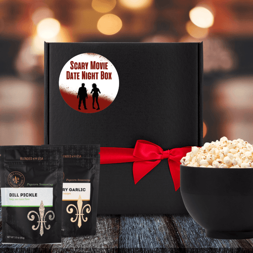 Black gift box with white label. Blood spilled and splattered on bottom half of label and a little across the top. The words Scary Movie Date Night Box in red are between the blood. Label shows a boy-girl zombie couple silhouette. Box is tied closed with red ribbon. Next to box is 2 popcorn seasoning pouches and a large bowl filled with popcorn. Dell Cove Spices
