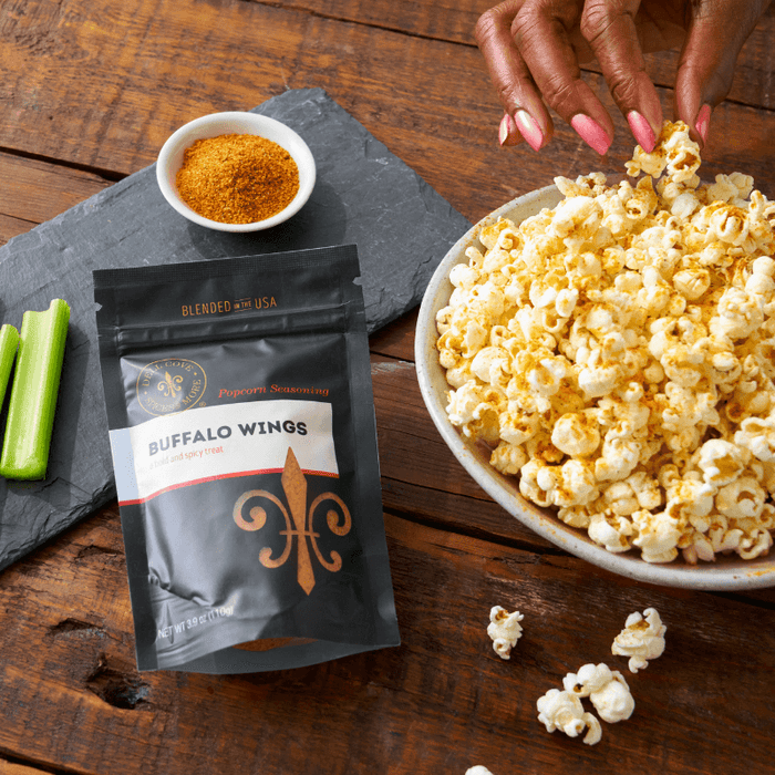Buffalo wings seasoning pouch on wooden table next to bowl of popcorn. Also, celery and a small bowl with seasoning. Hand is reaching for popcorn. Dell Cove Spices
