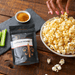 Buffalo wings seasoning pouch on wooden table next to bowl of popcorn. Also, celery and a small bowl with seasoning. Hand is reaching for popcorn. Dell Cove Spices