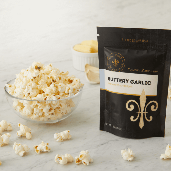 Buttery Garlic seasoning pouch next to glass bowl with popped popcorn and small bowl in background with butter and garlic cloves. Dell Cove Spices