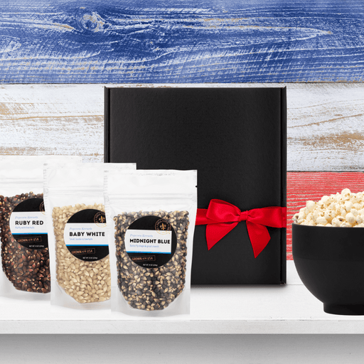 Black gift box tied closed with red ribbon next to 3 half pound popcorn bags, Ruby Red, Baby White and Midnight Blue with large black bowl filled with popped popcorn on red white and blue background - Dell Cove Spices