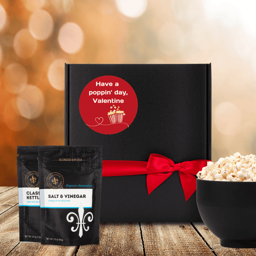 Black gift box with round red label that reads have a poppin' day Valentine with an image of two popcorn boxes and a white heart. Gift box is next to two popcorn seasoning pouches and a bowl of popped popcorn. Dell Cove Spices
