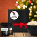 Black gift box with round white label featuring a red and green mini Christmas tree background, movie tickets, a movie clapper with a Santa hat and the words Christmas Movie Night. Box is next to two seasoning pouches and a large bowl of popcorn. Dell Cove Spices