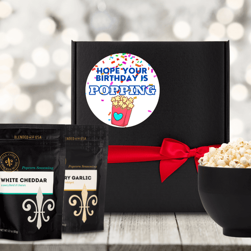 Black gift box featuring circle label with popcorn and confetti and Hop your Birthday is Popping text, tied closed with a red ribbon, next to a large black bowl filled with popped popcorn and 2 seasoning pouches- Dell Cove Spices