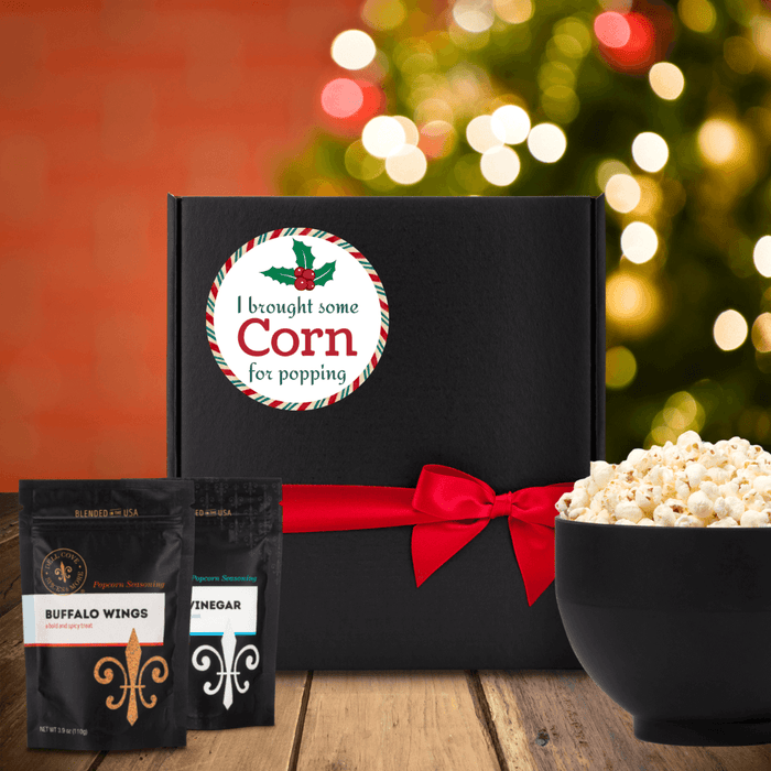 Black gift box with white round label that has red, green and white candy stripped edge, green holly at top and the words I brought some corn for popping. Box is next to two seasoning pouched and large bowl of popcorn. Dell Cove Spices