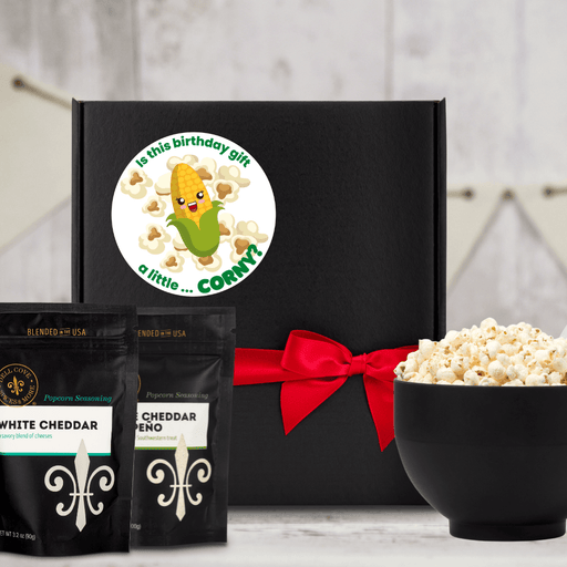 Black gift box featuring label with image of popped popcorn and ear of corn and text Is This Birthday Gift a little Corny, gift box is tried closed with a red ribbon. Next to large black bowl filled with popped popcorn and two seasoning pouches - Dell Cove Spices