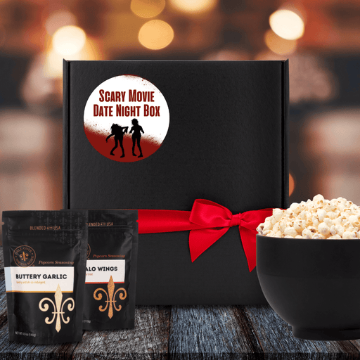Black gift box with white label. Blood spilled and splattered on bottom half of label and a little across the top. The words Scary Movie Date Night Box in red are between the blood. Label shows a girl-girl zombie couple silhouette. Box is tied closed with red ribbon. Next to box is 2 popcorn seasoning pouches and a large bowl filled with popcorn. Dell Cove Spices