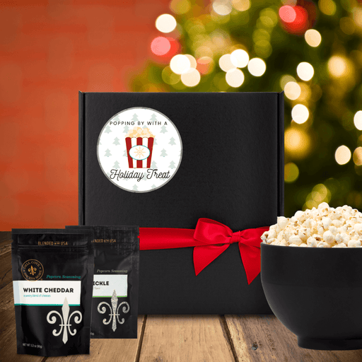 Black gift box with round white label, that features a background of mini green Christmas trees, an image of a red and white popcorn container with a gold snowflake on the front and the words popping by with a holiday treat. Box is next to two seasoning pouches and a large bowl of popcorn. Dell Cove Spices