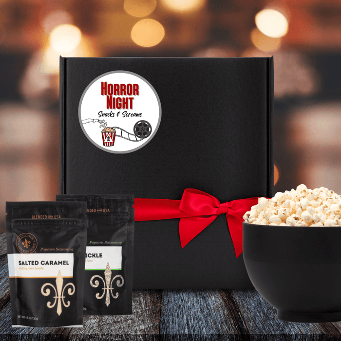 Black gift box with white round label featuring old fashioned movie reel and popcorn container will poison label. A skeleton hand is reaching for popcorn. The label also features the words Horror Night in red and snacks and screams in black. Box is tied closed with red ribbon. Next to box is 2 popcorn seasoning pouches and a large bowl filled with popcorn. Dell Cove Spices