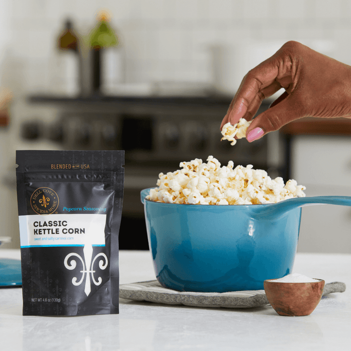 Classic kettle corn seasoning pouch on counter next to blue kettle filled with popped popcorn and small bowl with seasoning. Hand reaching over kettle, holding some popcorn. Dell Cove Spices