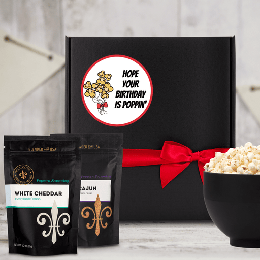 Black gift box featuring circle label with popcorn balloon bouquet and Hope Your Birthday is Poppin' text, tied closed with red ribbon, next to large black bowl filled with popped popcorn and 2 seasoning pouches- Dell Cove Spices
