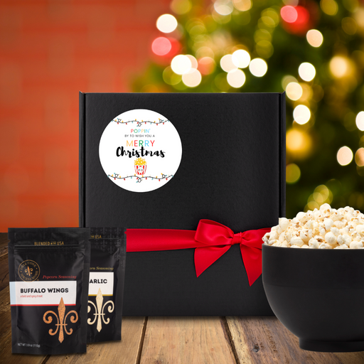 Black gift box with white round label, featuring strings of Christmas lights, a small cartoon popcorn container and the words Poppin by to wish you a Merry Christmas. Box is next to two seasoning pouches and a large bowl of popcorn. Dell Cove Spices
