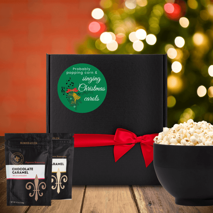 Black gift box with green round label. Label shows Christmas bell image with music notes and the words probably popping corn & singing Christmas carols. Box is next to two seasoning pouches and large popcorn bowl. Dell Cove Spices