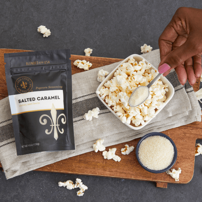 Salted Caramel seasoning pouch on wooden serving tray with a hand sprinkling seasoning from a spoon onto a small bowl of popped popcorn. Dell Cove Spices