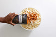 Large bowl of popcorn on white table with a hand reaching over and sprinkling seasoning from a Spicy Cajun seasoning pouch. Dell Cove Spices