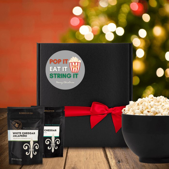 Black gift box with round gray label. Label has image of red and white popcorn container and the words pop it east string it in red, white and green and Merry Christmas in white. Box is next to two seasoning pouches and a large popcorn bowl. Dell Cove Spices