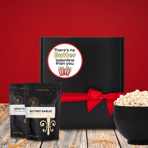 Black gift box with round label that reads there&#39;s no butter Valentine than you with an image of two popcorn boxes. Gift box is next to two popcorn seasoning pouches and a bowl of popped popcorn. Dell Cove Spices