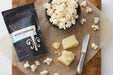 Small black bowl of popped popcorn next to White Cheddar seasoning pouch and block of cheddar cheese - Dell Cove Spices